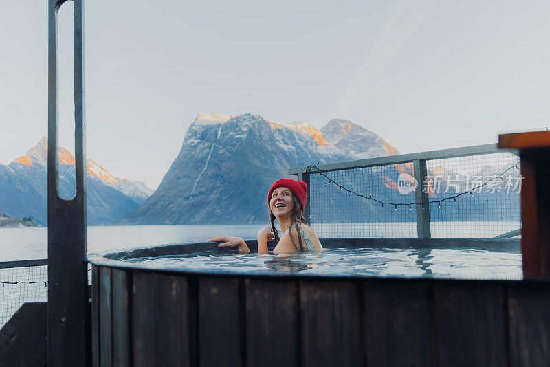 在挪威的冬季峡湾，一个快乐的女人在热水浴缸放松的红帽子的侧面视图