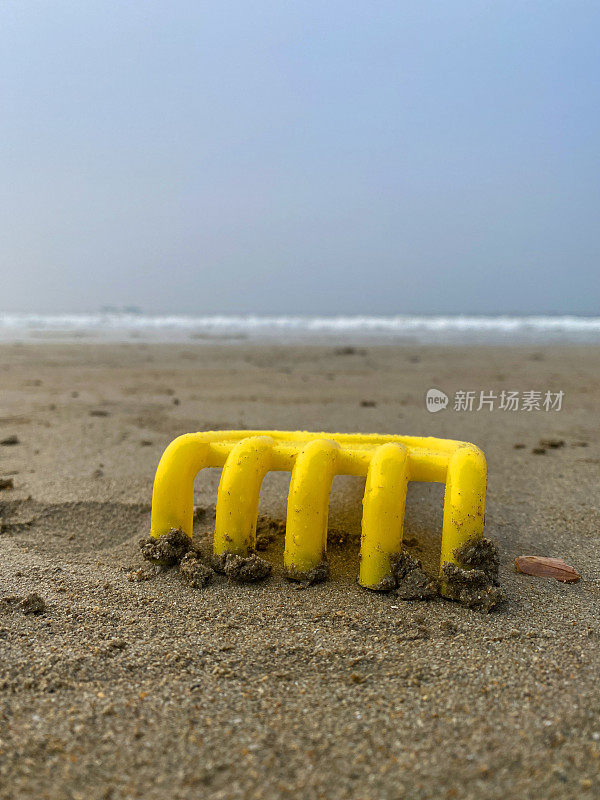 在沙滩上的塑料黄色耙子的特写图像，在退潮前的大海上鲜艳的儿童海滩玩具，关注前景
