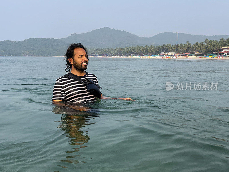 印度果阿帕洛莱姆海滩，一名印度男子在海滩度假，穿着黑白相间的t恤，胸前绑着防水袋，波浪起伏的水面上