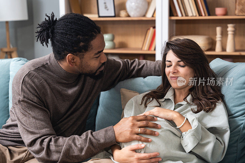 年轻男子未来的父亲帮助和协助他怀孕的女友未来的母亲在她怀孕。夫妇俩坐在家里的沙发上，丈夫安慰着抱着孩子的紧张的妻子。