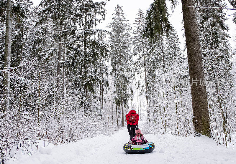 一名年轻女子带着一个8岁的孩子走在路上，她的孩子坐在一个充气环上。一家人在下雪的公园里散步