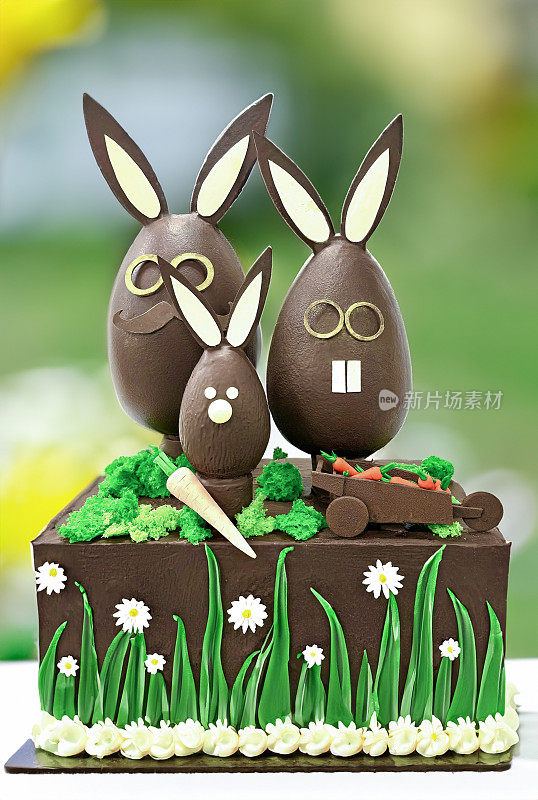 三个大型的，模塑的，牛奶巧克力复活节彩蛋兔子站在巧克力花园设计的展台上，巧克力小推车的糖霜胡萝卜，雏菊花装饰，模糊了春天花园的背景
