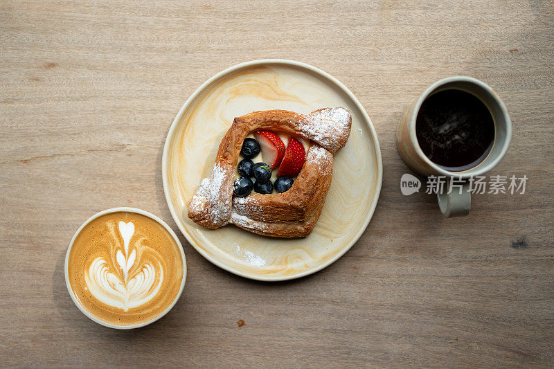 英式早餐，有混合浆果、丹麦面包、美式咖啡和咖啡拿铁，放在木桌上。