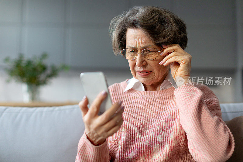 困惑的戴眼镜的老年妇女眯着眼睛看手机，视力有问题，坐在沙发上