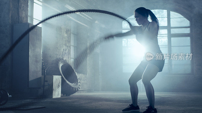 强壮的女运动员练习与战斗绳索作为她的交叉健身健身房锻炼的一部分。她满身的汗水和训练发生在一个废弃的工厂改造成健身房。