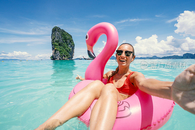 快乐的女人在泰国岛屿清澈的海水中与粉红色的充气火烈鸟在田园般的海滩上自拍。人们旅行奢侈，乐趣和酷的态度概念