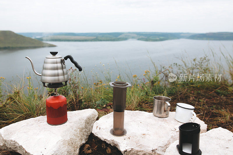 钢水壶沸腾的天然气primus，金属杯子和气压在悬崖上的背景湖，冲泡咖啡在露营。在户外野餐时制作热饮。在山上徒步旅行和远足