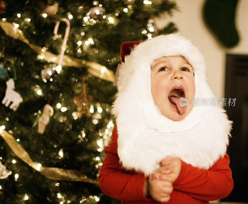 一个三岁的白种人男孩戴着圣诞老人帽和胡子在圣诞树前调皮地伸出舌头