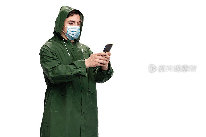 年轻男性戴着N95口罩，穿着绿色雨衣，在白色背景下使用智能手机