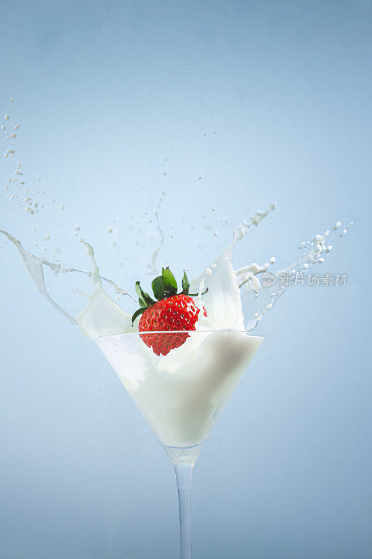 草莓滴入牛奶鸡尾酒杯，在蓝色背景上产生飞溅。