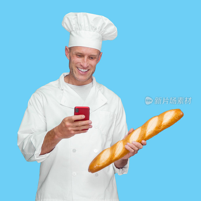 白色的年轻男性面包师前面的蓝色背景穿着裤子，拿着一条面包，并使用短信
