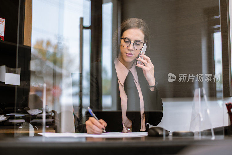 戴眼镜的专注年轻女商人坐在办公桌前用手机聊天写文书文件，专注年轻女商人用智能手机与客户谈合同