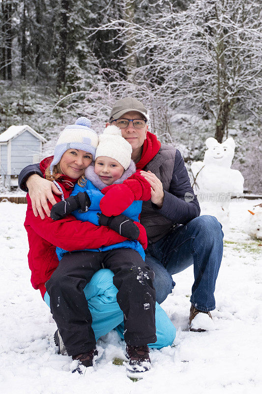 一个有孩子的家庭，一个五岁的白人女孩。一家人拥抱着，看着镜头，背景是一个雪人和森林。