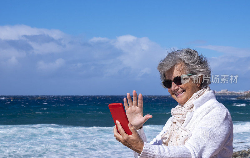 有吸引力的老年妇女与灰色头发享受海滩度假在一个大风天使用智能手机视频通话。轻松微笑活跃的退休人员