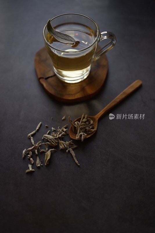 鼠尾草茶在玻璃杯与柄木制杯垫在黑色混凝土背景。有一片新鲜的鼠尾草茶叶放在鼠尾草茶上。杯子旁边是一个木勺和撒在地上的干鼠尾草茶。花草茶。