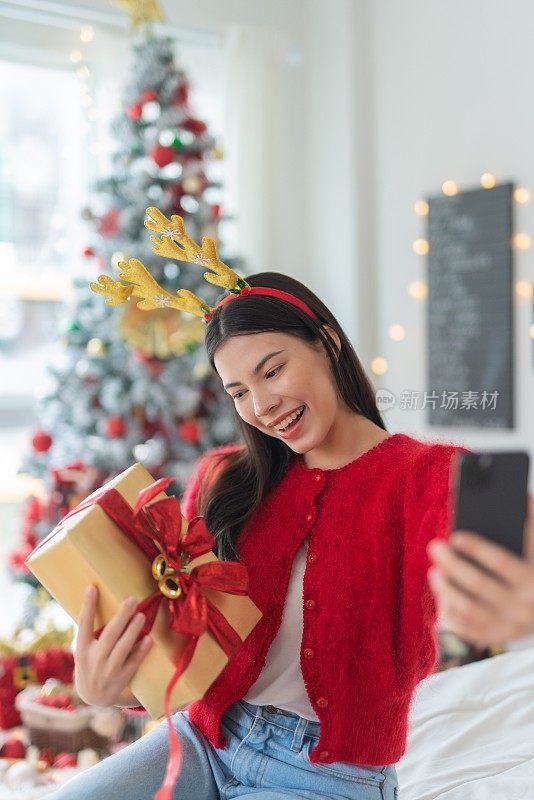 肖像照片的年轻美丽友好的亚洲女性与可爱的圣诞发带高高兴兴地拿着礼盒和自拍使用她的手机在一个漂亮的装饰圣诞树作为背景