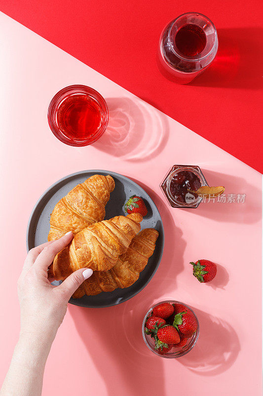 一个女人的手拿着一个kraussant放在一个盘子上，盘子里有新鲜的牛角面包、浆果汁、果酱和新鲜的浆果，背景是粉红色和红色的。