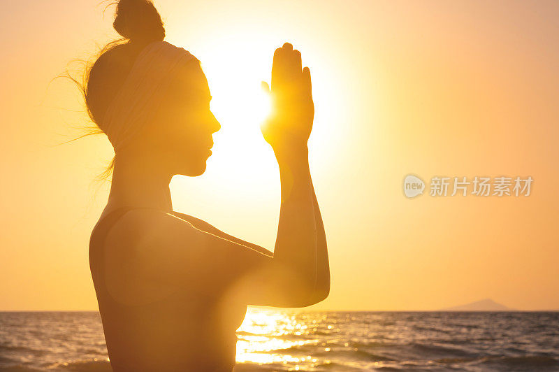 一个女人的剪影坐在岸边，双手合十祈祷。日落或黎明时分，在海边交叉双手进行精神活动的妇女