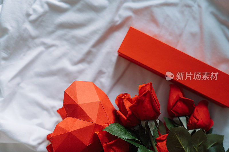 纸红色的心，上面有一份礼物和一束玫瑰，放在一张皱巴巴的白色床上