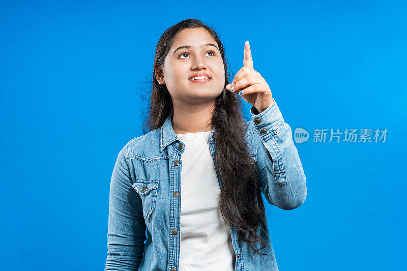一个迷人的开朗的亚洲女孩的肖像，在蓝色的背景上用食指向上指示
