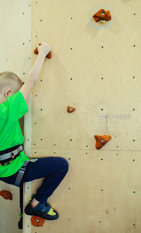 儿童攀岩。穿绿衬衫的金发男孩正在爬墙。抱石类。为安全活动及比赛而设的人造跑道。登山，安全系统，专用鞋