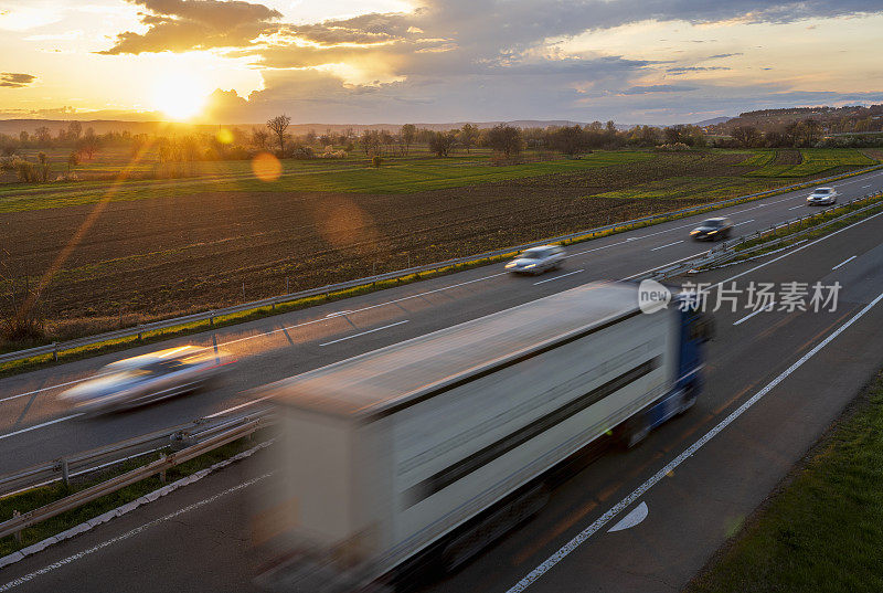 一辆大卡车和几辆汽车在公路上高速行驶，穿过乡村风景。高速公路高速行驶。高速公路上超速行驶的景象。背景是美丽的日落。背景是美丽的日落。