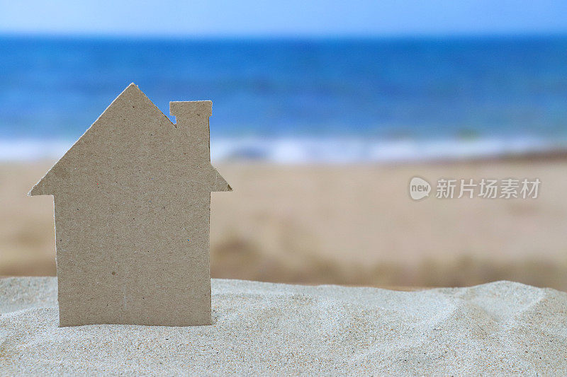 在一堆沙子上的纸板剪纸模型度假小屋的特写图像，沙滩背景与退潮时的波浪，大海和海岸线，重点放在前景，度假租赁和度假屋的概念，复制空间