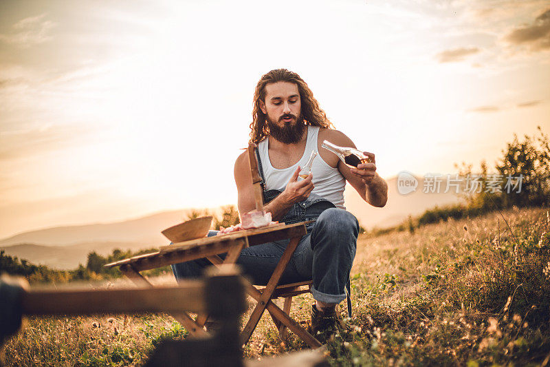 自然的守护者。一个在山上的男人，一个伐木工人，喝着自制的白兰地，吃着自制的食物，而在他身后是美丽的日落，休息辛苦的工作。