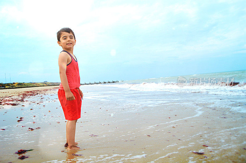 一个小男孩穿着休闲的衣服站在沙滩上，在海浪中摆姿势