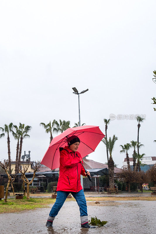 一个女人在雨天打着红伞走在公园里。一个穿着靴子的模特踩在水坑上