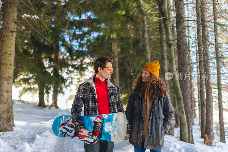 一对美丽的年轻异性恋情侣在雪原森林的小路上散步，手里拿着一块滑雪板