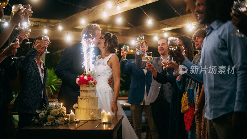 美丽的新娘和新郎在晚餐会上与多民族朋友庆祝婚礼。新婚夫妇提议为幸福婚姻干杯，站在有蛋糕和烟火的餐桌旁。