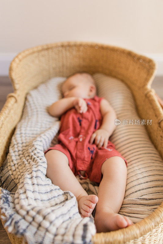 一个昏昏欲睡的18周大的男婴穿着红色航海服躺在一个舒适的奶油色条纹棉毯在海草摩西篮子