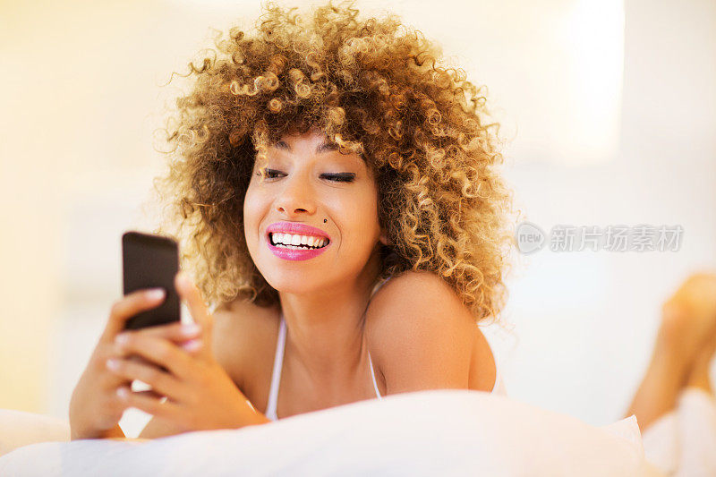 微笑的非洲裔美国妇女在床上使用智能手机。
