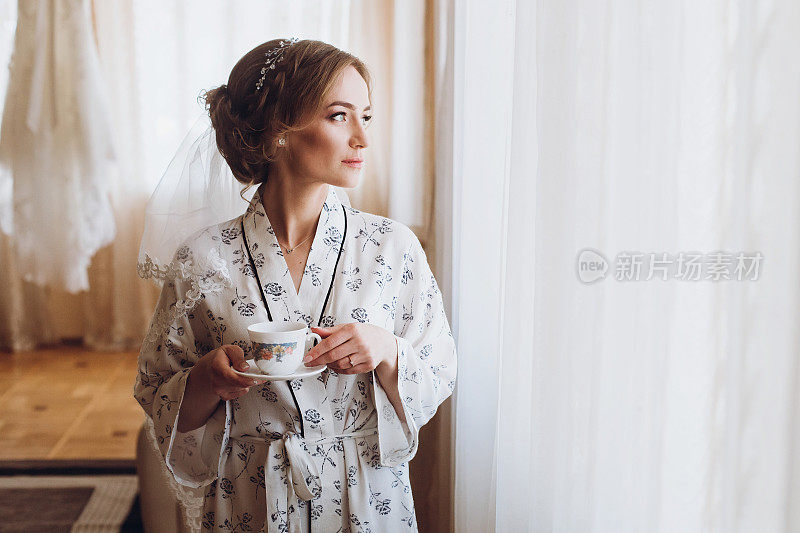 华丽的新娘穿着优雅的长袍，在豪华酒店靠近窗户的房间喝咖啡，早上婚礼前的准备
