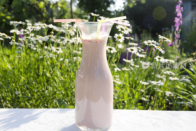 美味的草莓奶昔。鲜花草地上的桌上放着一瓶粉红色的饮料和吸管