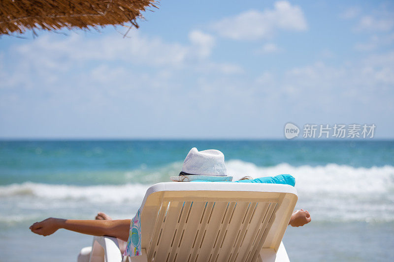 难以辨认的斜靠在一个白色帽子的女人在海滩上晒太阳，以大海为背景，特写
