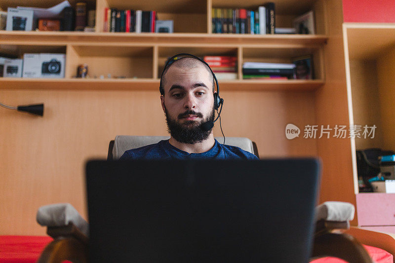 秃顶的年轻人戴着带麦克风的耳机在家里用笔记本电脑工作