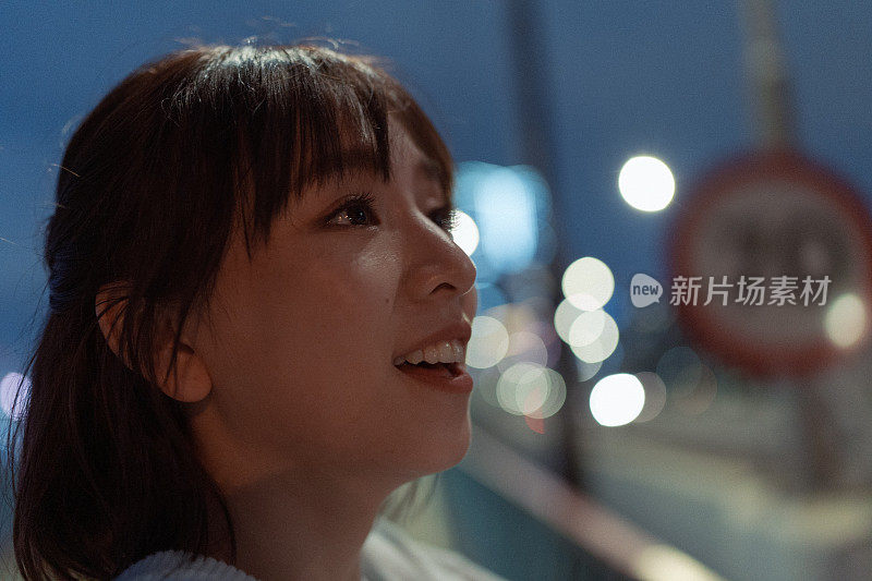 特写的年轻微笑的亚洲妇女的脸看出去和远离夜景与城市的灯光在街道上