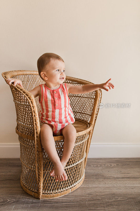 2023年，南佛罗里达，一个可爱快乐的17个月大的小男孩，浅棕色的头发和棕色的眼睛，穿着红色和奶油色的条纹纱布连衣裤，赤脚坐在一张老式藤椅上。男婴有轴后多趾畸形
