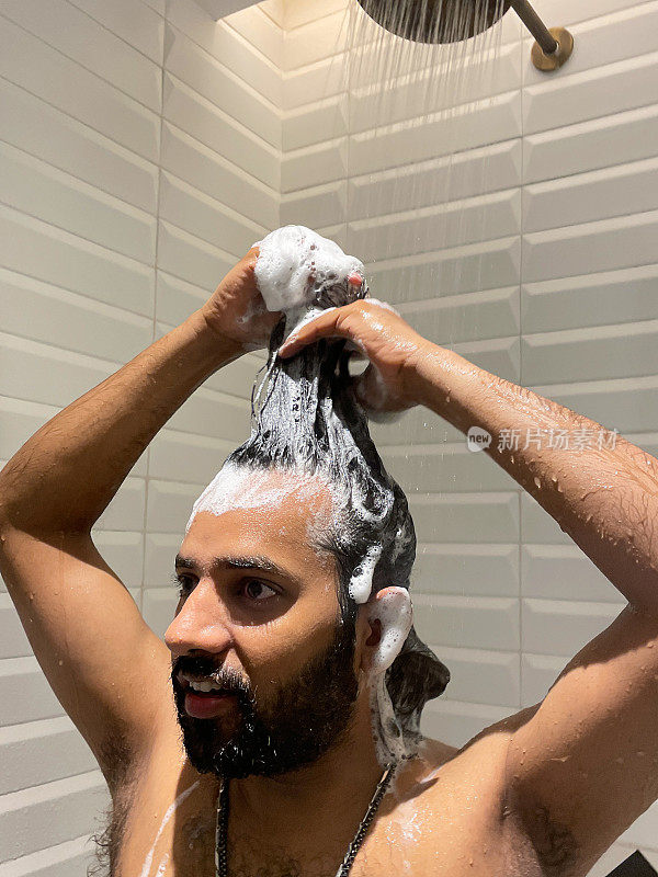 印度男子在淋浴间洗头的特写图像，使洗发水泡沫在流水下卷曲，白色斜边墙砖水平堆叠图案，重点放在前景