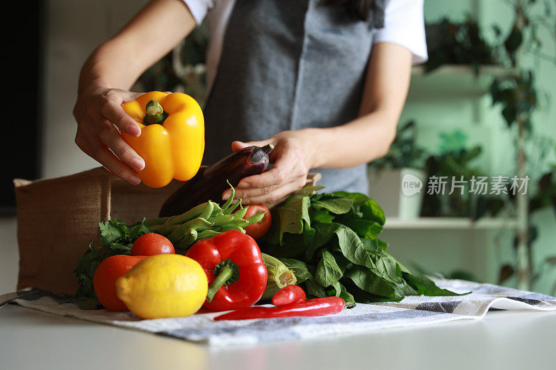 一位年轻的亚洲妇女从杂货店购物回家，从可重复使用的袋子里取出新鲜健康的有机水果和蔬菜放在桌子上。这反映了负责任的购物、零浪费和可持续的生活方式理念。