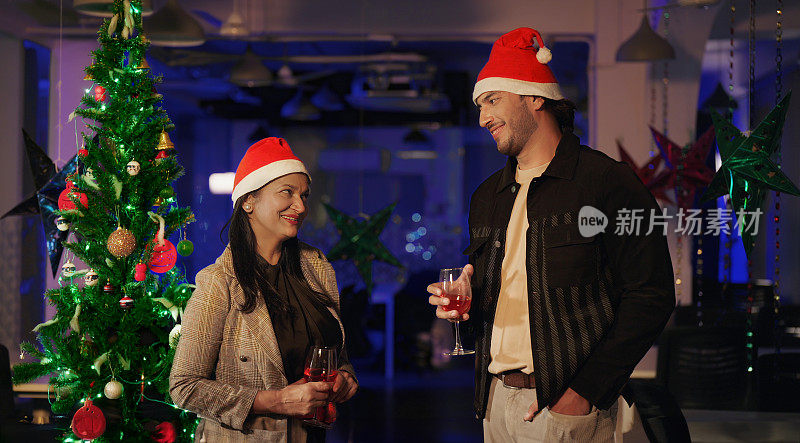 两位戴着圣诞帽的印度同事站在室内办公室，端着杯子，喝着果汁，一起享受夜晚的2024年新年晚会。快乐微笑的漂亮女性年轻男性做有趣的八卦