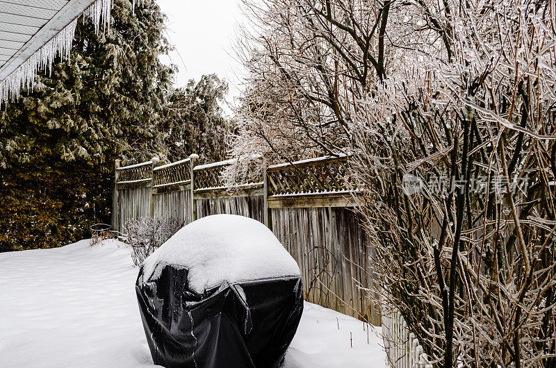 冬天郊区的后院有一个被雪覆盖的烧烤架和被冰覆盖的树枝，冰柱从冻雨。