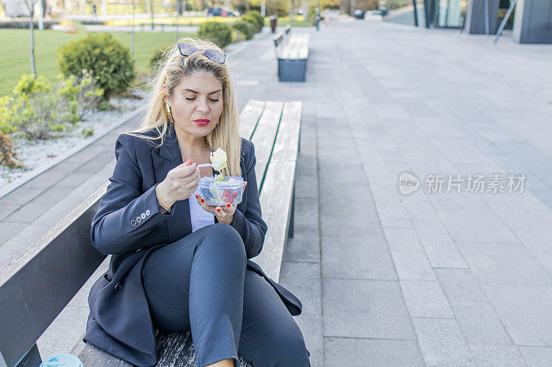 在户外的午餐休息时间，一位微笑的女商人正在吃健康的沙拉。在户外灵活工作。人们在使用科技的同时享受城市的绿色空间。