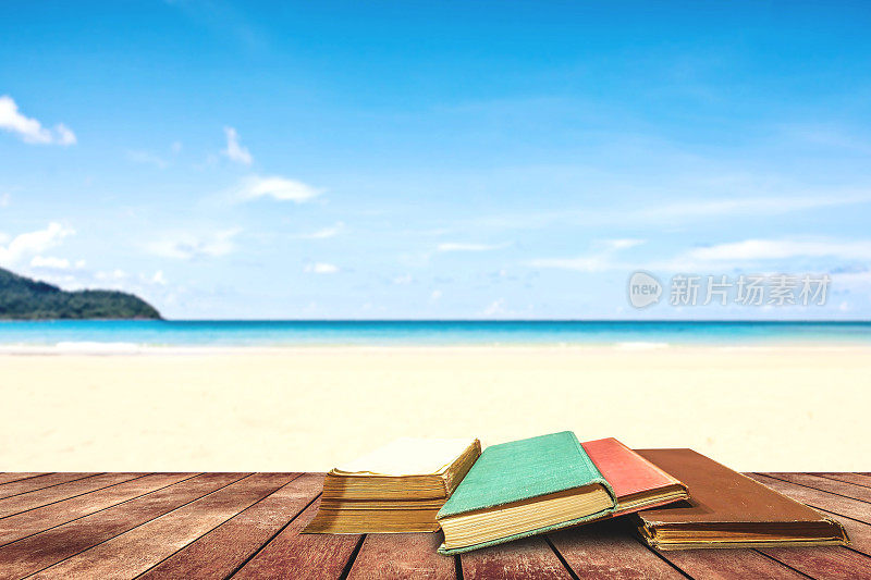 在白天正午的阳光下，热带海滩和蓝色的大海旁边的木制平台上，放着树木老化的书和垃圾纸。