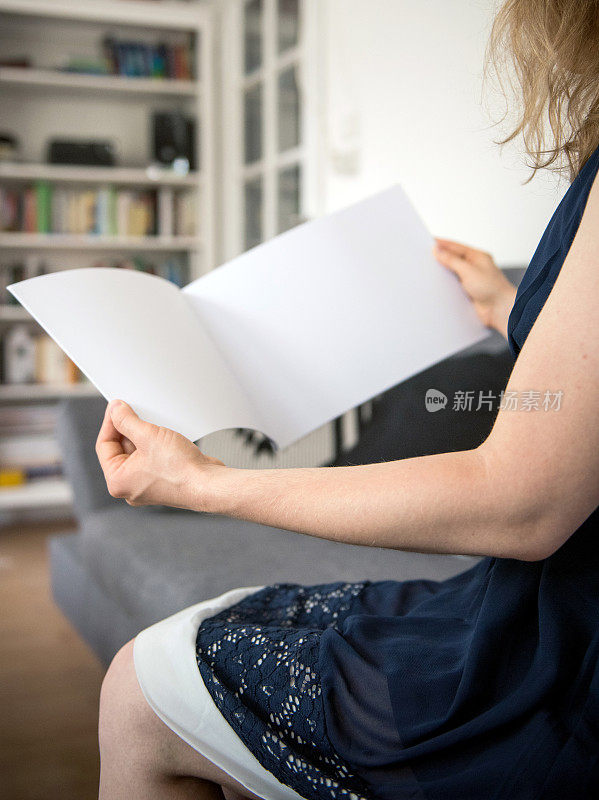 年轻女子坐在家里的沙发上，正在阅读和打开一个空白的白纸小册子副本空间