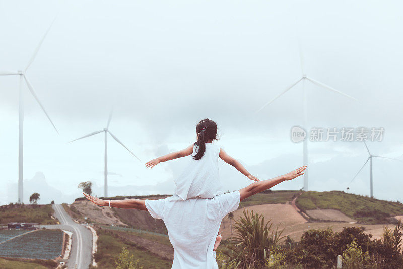 亚洲小女孩骑在父亲的肩膀上，一起在风力涡轮机场玩的背影
