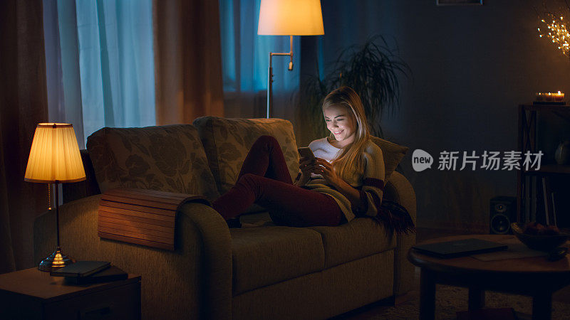 晚上，美丽的年轻女子躺在沙发上使用智能手机。房间看起来温暖舒适。