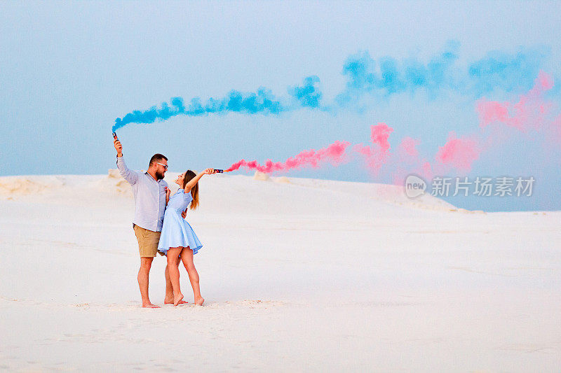 一对年轻的情侣站在沙滩上，手里拿着烟雾弹，看着对方，浪漫的情侣在沙滩上拿着蓝色和红色的烟雾弹